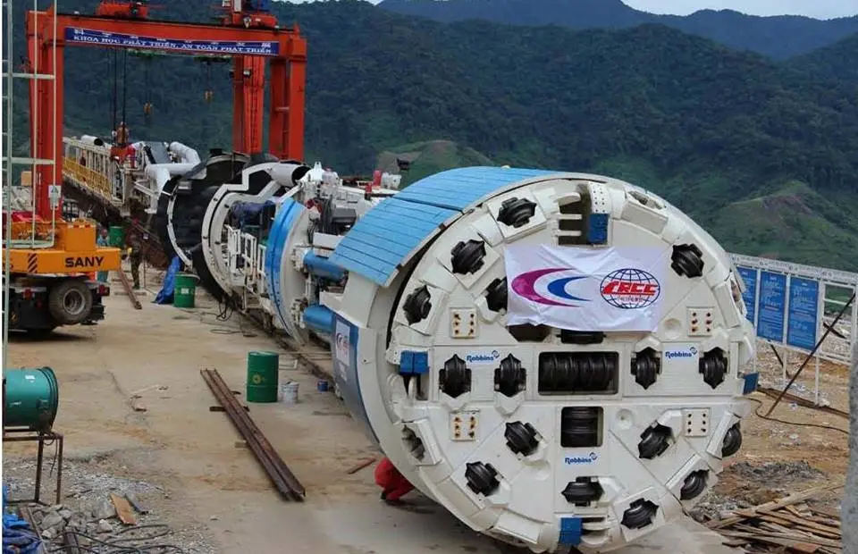 Máy đào hầm TBM hiệu Robbins đặt trên công trường, đầu cắt hình tròn gắn nhiều đĩa cắt, phía sau là cần trục cổng màu đỏ và núi rừng.