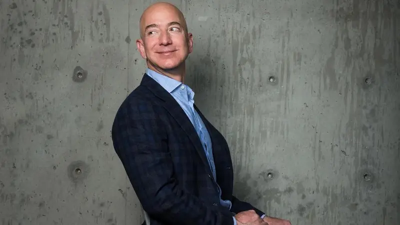 Jeff Bezos mặc vest kẻ ô tựa lưng vào bức tường bê tông