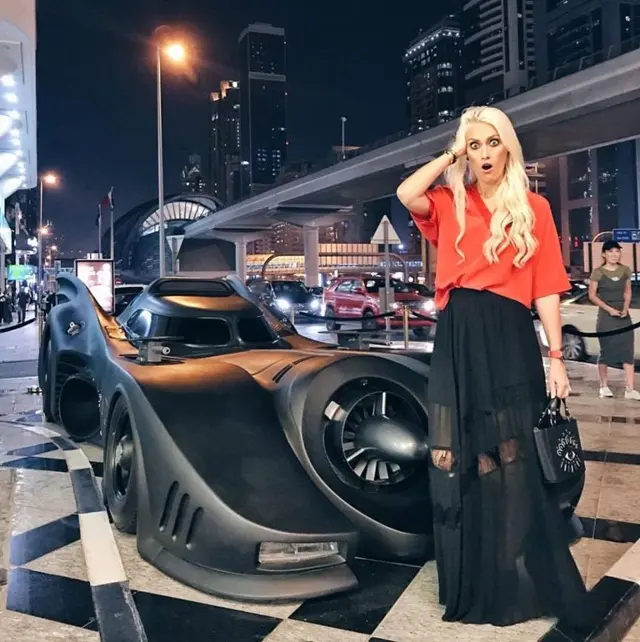 Người phụ nữ tóc vàng đứng cạnh xe Batmobile màu đen