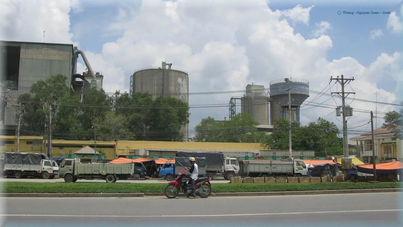Nhà máy xi măng với các silo, xe tải đậu dọc đường phía trước