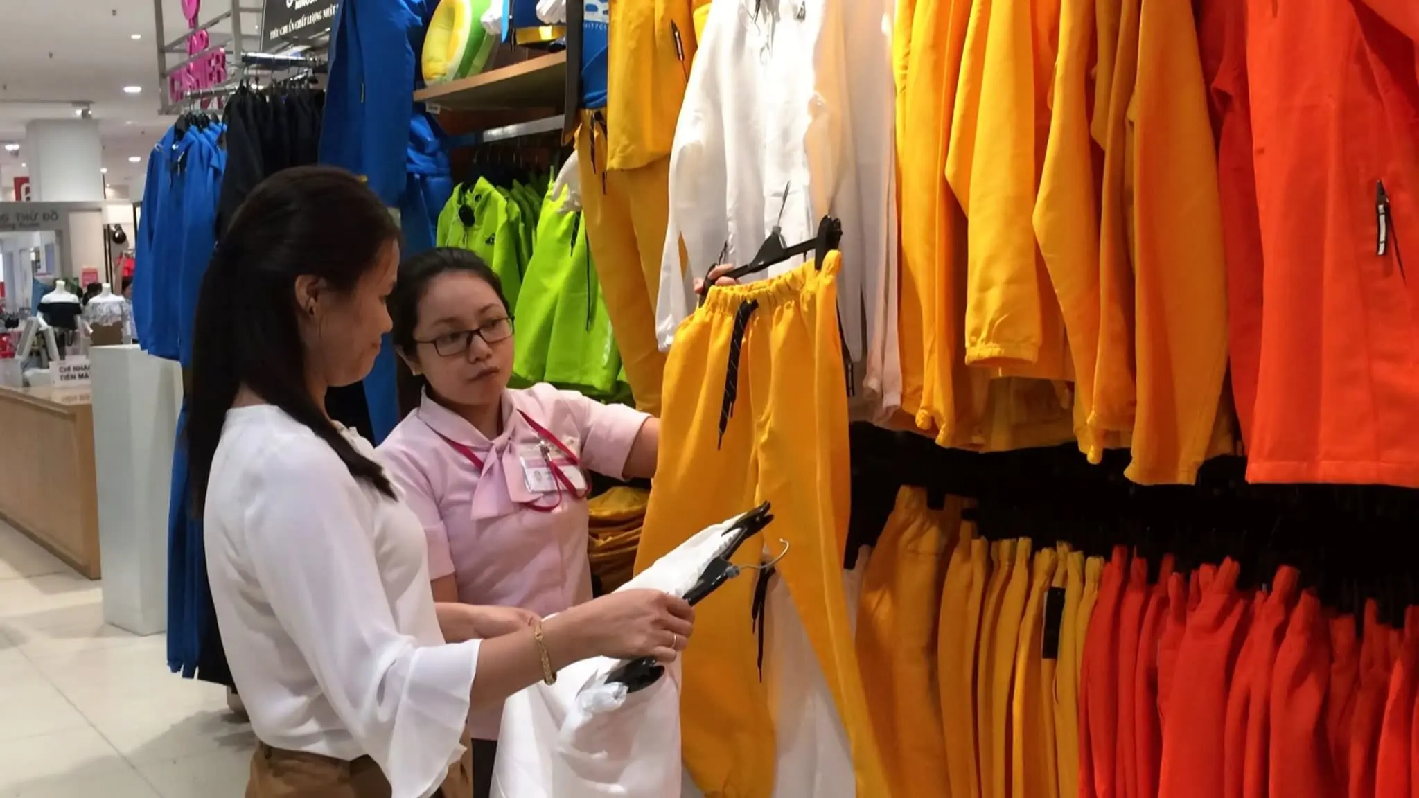 Nhân viên áo hồng tư vấn khách xem quần áo nhiều màu trên giá